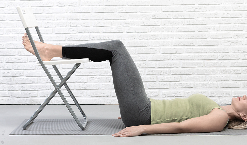 Kobieta leżąca na macie do jogi na podłodze z nogami uniesionymi na krześle równolegle do tułowia