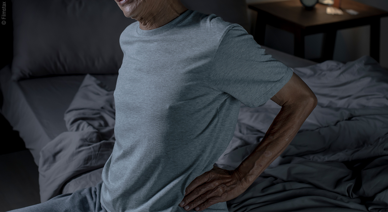 Osoba siedząca na łóżku, chwytająca się za plecy z powodu bólu